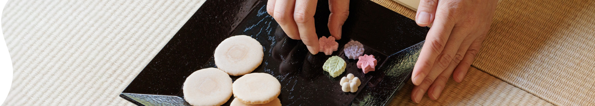 和菓子についてのページタイトルの背景画像。四角形の黒い皿の上に落雁とせんべいが乗っていて、落雁を手で持っている画像。
